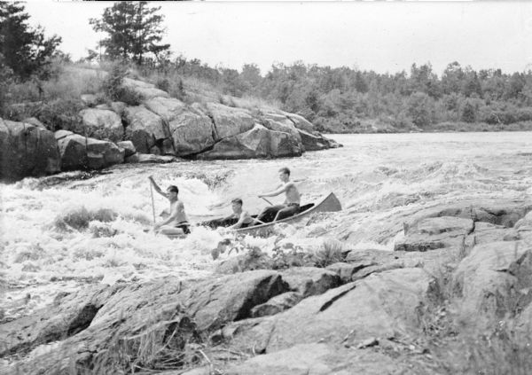 View from rocky shoreline towards three young men in a canoe running the Little Falls rapids on the South Fork of the Flambeau River.  