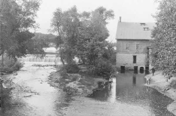 Elevated view of a man standing on a bank on the right, and two boys standing on a log on the left, are fishing in the mill stream below an old mill. Two boys without fishing poles are watching from the bank near the man. Water is flowing over the dam on the left.