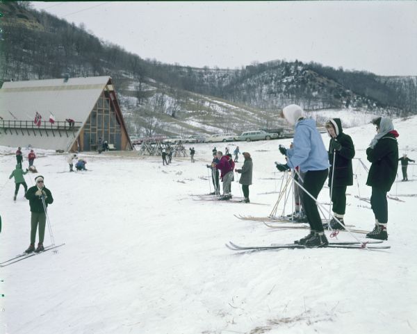 A group of women skiers, one wearing ski boots but no skis, are standing on the slope of a hill just above an A-frame lodge. Cars are parked in  the parking lot, and other skiers are nearby. Two people are standing on a balcony built along the side of the roof of the lodge. Several flags are on flagpoles on the balcony, including those of Norway and the United States. There is light snow cover on the surrounding hills.