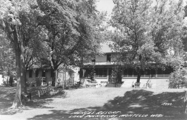 Photographic postcard view of trees shading the lawn at Menges Resort on Lake Puckaway. There is a two-story wood frame building with a screened porch across the front and a balcony above. There is a cottage to the left, and a dog and a man are in the yard in front. Steps with a rustic log railing lead up the sloped lawn to an arbor. A hammock is hanging between two trees in the foreground on the left, and there are outdoor benches near the porch. On the far right in the foreground is an archery target set up on a stand. Caption reads: "Menges Resort, Lake Puckaway, Wis."