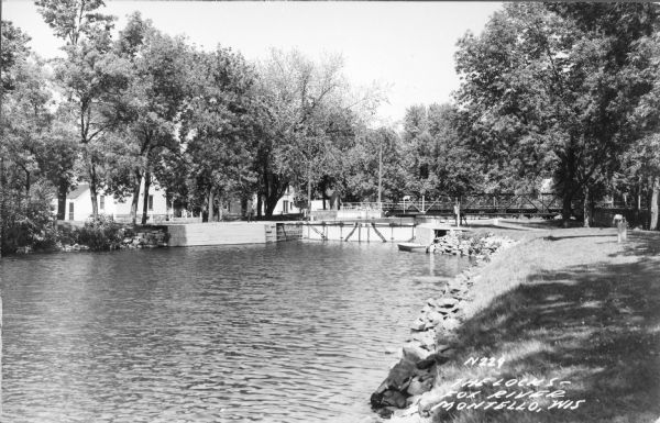 Photographic postcard view from shoreline looking downstream at the Fox River locks, with a bridge behind it. A boat is tied along the shoreline just below the docks. Two houses, facing each other across Main Street, are partially obscured by trees on the left. Caption reads: "The Locks, Fox River, Montello, Wis."