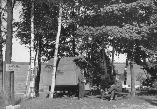A woman wearing a bandana on her head is sitting at a picnic table talking with another woman who is standing in front of a tent-style camping trailer. Both women are wearing plaid shirts and jeans. Birch and other trees are shading the campsite. There is an automobile on the right with the trunk lid propped open. Swimming suits and towels are drying on a line strung between two trees. Sturgeon Bay is in the background. A sign on the far left reads: "Camp Area No Picnicking."