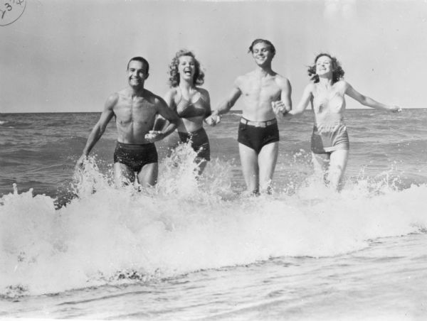 View from shoreline towards two men and two women in bathing suits standing and holding hands while standing in shallow water as a small wave breaks around them. On the reverse of the photograph is written: "Bathers at a Wisconsin beach on Lake Michigan."