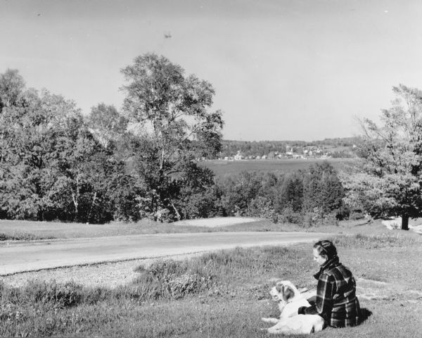 View looking down towards a woman wearing a plaid jacket sitting on the ground with her hand on a dog lying beside her in Peninsula State Park. There is a narrow road in front of them with a wooded area on the other side. The village of Ephraim is in the far background beyond the waters of Eagle Harbor.  