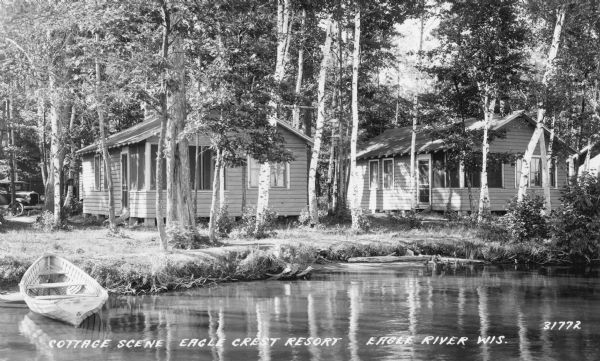 Photographic postcard view across water towards two waterfront cottages, shaded by birch and other trees. There is a wooden canoe pulled up to the shore on the left. The cottages feature small, enclosed screened porches, and an automobile is parked under the trees behind the cottage on the left. Caption reads: "Cottage Scene, Eagle Crest Resort, Eagle River, Wis."