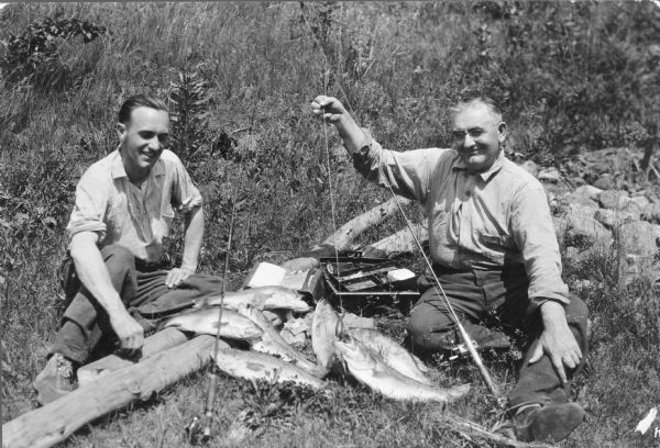 Two smiling men are sitting on the ground and posing with seven good-sized fish. The man on the right is holding a line with a lure on it above the open mouth of one of the fish. There is a tackle box on the ground in the center, and two rods with reels are propped in the foreground.