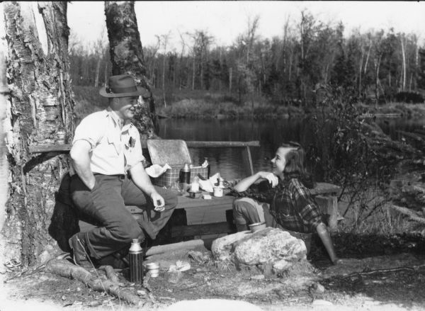 A man, sitting on a wooden bench, and a woman, sitting on the ground, are enjoying a picnic on the shore of the Flambeau River. There is an open plaid metal picnic hamper, utensils and thermos bottles nearby. The man is holding a cigarette.  