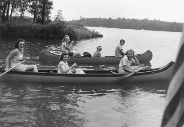 View across water towards six women, three in each of two canoes, are smiling as they paddle near a shoreline in the background. There are buoys farther out in the water in the background on the right, and the far shoreline is in the distance. In the right foreground is part of a United States flag.