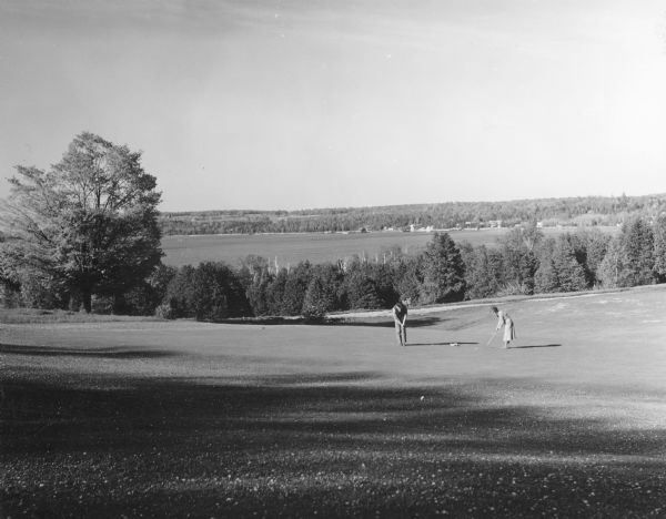 View down hill towards a man and a woman posing in the act of putting on a green of the golf course in Peninsula State Park. Eagle Harbor and the village of Ephraim are in the background across the bay.