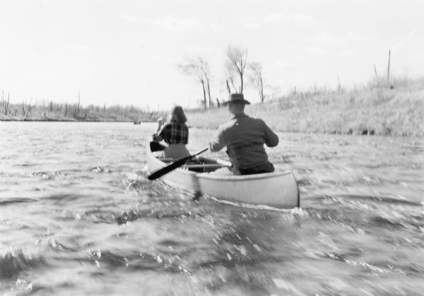 An unidentified man and woman are paddling their canoe against the current in the fast running Flambeau River. A small stand of leafless trees and blackened tree trunks on the banks provide evidence of a prior forest fire.