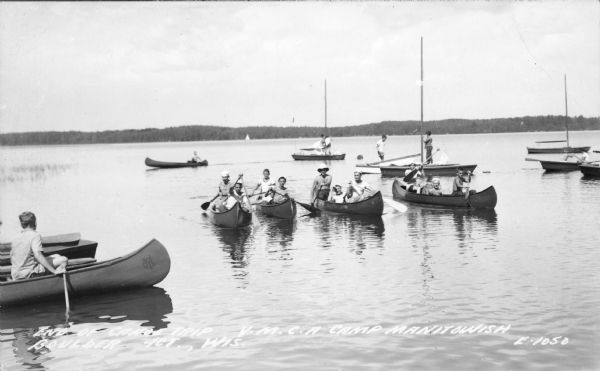 Photographic postcard view of young men and boys paddling canoes toward the shore of Boulder Lake. A canoe on the left bears the YMCA insignia.  Other youth are working on small sailboats in the background. C aption reads: "End of Canoe Trip Y.M.C.A. Camp Manitowish [sic] Boulder Jct., Wis." 
