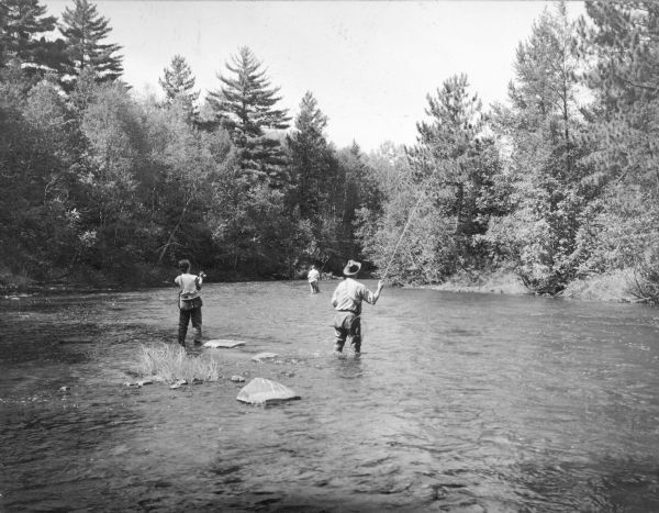 Three anglers wearing waders, including a woman on the left, are casting while standing in shallow water. The woman has a kreel on her hip, and the man on the right is carrying a net. A typed caption on the reverse of the print reads: "How's this for an ideal spot for fly fishing? A river in Burnett county, northwestern Wisconsin." 