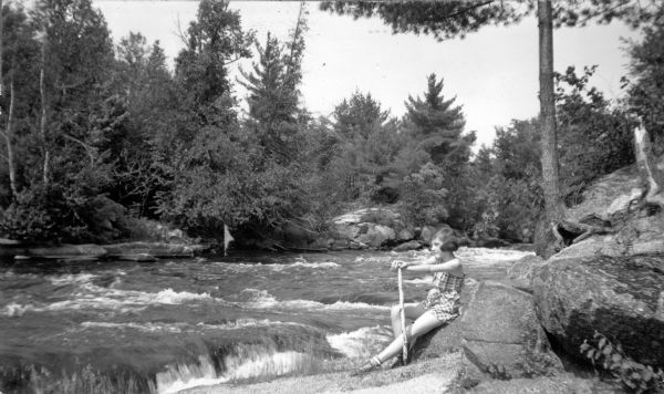 A young woman is posing sitting on a boulder on the shoreline along a river rapids. She is holding a walking stick and is wearing a romper. The description on the reverse of the photograph reads: "An inviting spot to rest and watch the fast waters of the Wolf River flowing thru [sic] the wild forest region of the Menominee Indian Reservation, Shawano County, Wisconsin."