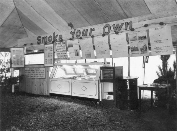 A display by the Wisconsin Conservation Department erected in a large tent extols the value of smoking fish for food. There are posters identifying different fish species and describing fish harvesting by seining. A sign overhead reads: "Smoke Your Own." Smoked fish are displayed in a case, center, with different smoking equipment set up at right. At far right, a man is writing at a desk obscured by a small pine tree.  