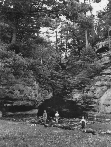 Two girls and a boy are posing standing at the edge of the pool at Pewit's Nest. The boy has rolled the legs of his overalls above his knees. There are tall rock outcroppings and trees behind the children.
