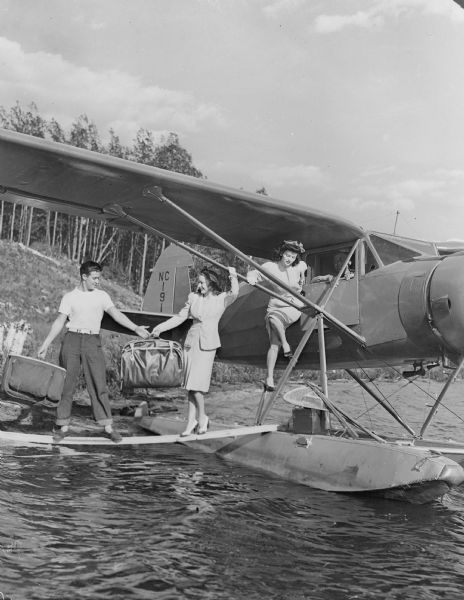 A young man wearing jeans and a t-shirt is handling the luggage as two well-dressed women exit a small seaplane. The pilot is watching from inside the plane. There is a single plank from the float to the shore, which rises to a wooded slope.