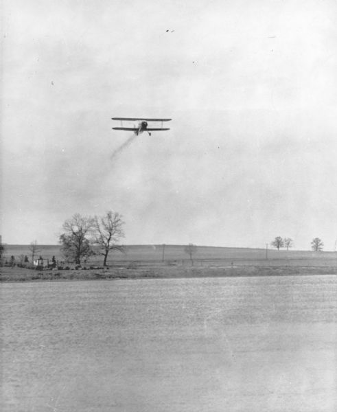 A biplane is dropping fingerling fish into a Wisconsin lake. There are two small sheds on the shoreline. The caption on the reverse of the print reads: "Fish have been successfully transported over long distances and planted from the air by the Fish Management Division of the Wisconsin Conservation Department. The method offers speed and flexibility in distribution of fish. Further studies are being made as to its practical application in this state. The photograph shows the fish leaving the airplane in an experimental planting test."