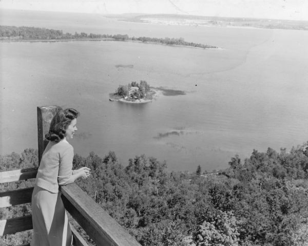 A young woman smiles as she enjoys the view from the Potawatomi Tower in Potawatomi State Park.  There is a house on a small island in Sawyer Harbor, with Cabot Point and northern Door County in the background. 