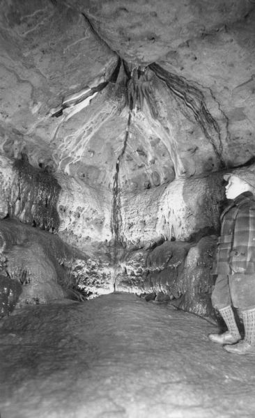 Arthur Pond, wearing knickers, heavy stockings, a coat and hat is standing admiring the walls in the Cave of the Mounds. On the reverse of the photograph is the caption: "Above the cave['s] onyx 'glacier' hang rare black and white ribbons of stone striped as evenly as if woven on a loom. These are found no where else in the world except in a cave in Iceland. The black stripes are colored by oxide of manganese which sometimes stain the lime-saturated water which trickled along the ceiling and dripped off to the ledge (left) forming the black knobs, Black Druids guarding the beauty of the cavern."