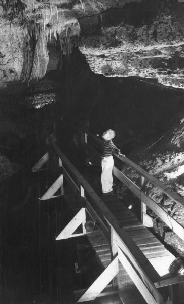 The typed caption on the reverse of this photograph explains: "On the bridge above the underground lake in Cave of the Mounds at Blue Mounds, Wis., Arthur Pond admires the Crystal Chandelier." Stalactites which have formed in rows and tiers create the chandelier.  