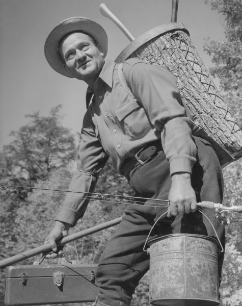 An unidentified man is posing outdoors wearing a straw hat, long-sleeved shirt and slacks, with a bark basket worn as a backpack. He is carrying a tackle box and bait bucket. There is an axe handle protruding from the basket. On the reverse of the photograph is written: "Fisherman with his camping outfit."