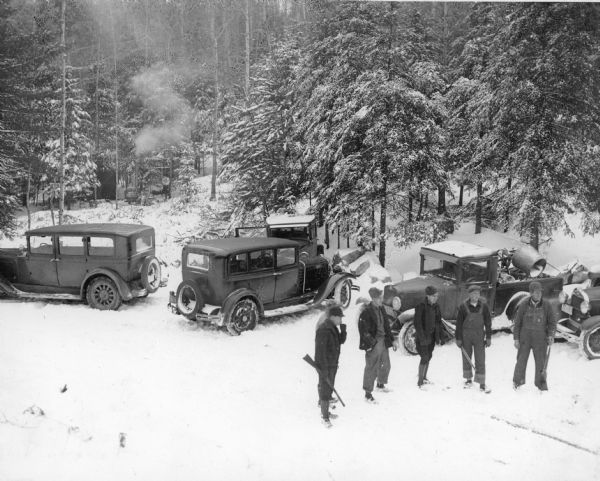 Elevated view of six hunters, one obscured, standing near four cars and a pickup truck parked at the edge of the woods. There is snow on the ground. Three of the hunters are holding guns, and several men are wearing bib overalls. There is a cement mixer in the back of the pickup. There is a tent on the left, obscured by trees. Smoke is rising from the flue of an outdoor stove near the tent. On the reverse of the photograph is written: "Eagle River Deer Season."
