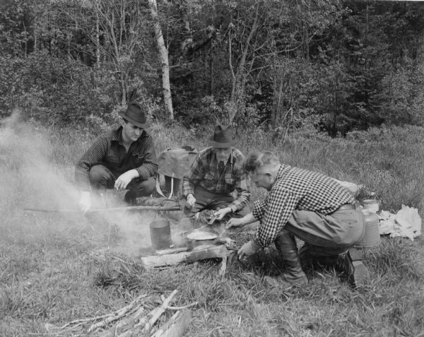 Three men are preparing supper over a small campfire at the edge of a wooded area. At right, two men are tending to food cooking in frying pans, while the third man is steadying a pole supporting a tin can over the fire. There is a large Thermos jug in the background. Two of the men are smoking cigarettes.  