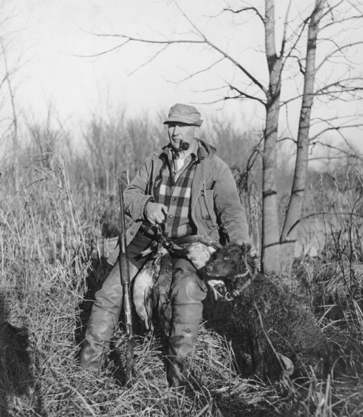 A man wearing waders and smoking a pipe, is sitting and holding several ducks on his lap, and has his shotgun resting on his right leg. His left hand is resting on the head of his hunting dog, probably a Curly-Coated Retriever.