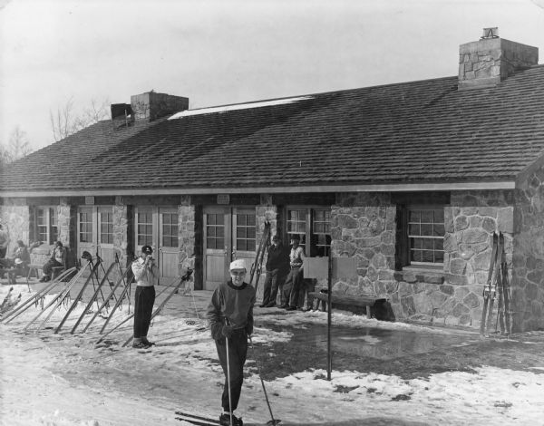 A man on skis and holding ski poles is smiling at the camera in front of the stone warming house at Rib Mountain State Park. Skis and poles are propped on stands, and other skiers are taking a break. Two men are leaning against the building and two women are sitting on a wooden bench on the left. The warming house has two large stone chimneys.  