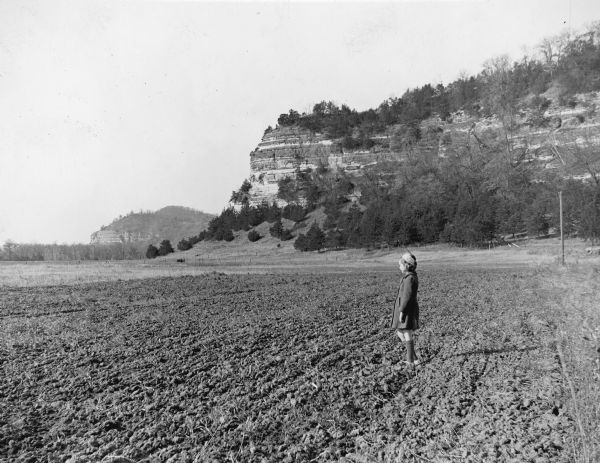 A young girl wearing a winter coat and hat is standing in a tilled field at the base of Cassell, or Leykauf, Bluff. A description on the reverse of the print states: "On [Highway] 60 to Spring Green." There is another bluff in the far distance.