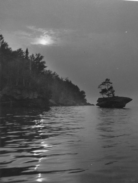 The moon, partially obstructed by clouds, is reflected in the waters off one of the Apostle Islands. There is a small islet on the right, with several trees growing on it.