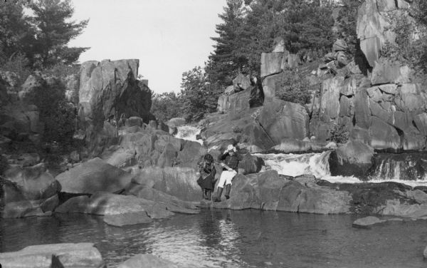 View across water towards an unidentified man and woman sitting on rocks at the edge of the Pike River, with Dave's Falls behind them.