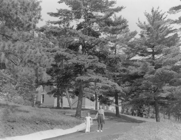 An unidentified woman and girl are posing on the drive leading to a brick house behind mature pines. The house was built in the 1890's by General Walter Cass Newberry as a summer home on the former estate of Governor Nelson Dewey. Dewey had built a house on this site in 1868, which burned in 1873. The Newberry house was built on the foundation of the old house, and is now part of Stonefield Historic Site.