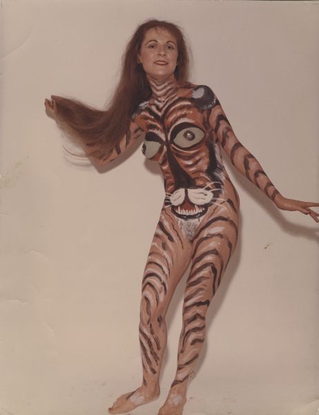 A nude woman, with long hair, is standing and posing against a backdrop. Her body has been painted to look like the face of a tiger, with stripes on her arms and legs. 