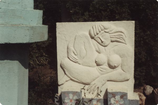 Finished large rectangular concrete-slab bas-relief of a nude woman identified as "Diana." She has long hair and bangs, and is sitting in a relaxed pose. Supported by her left elbow, her left leg wraps around the right at the ankle, with her right hand falling below the knee. The sculpture measures 45" x 41" x 14." The base of the slab is painted bluish gray with white columbine flowers. Part of a green sculpture is on the far left. 