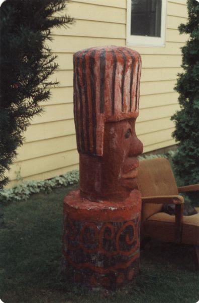 Side profile of a Primitivism-styled, cast-concrete sculpture of a male figure in Sid Boyum's backyard near yew bushes and the side of the house. The figure is wearing a red and dark blue striped headdress. The head is on a cylindrical base of the same two colors but decorated with curvilinear patterns and circles. On the right is an armchair with a sleeping cat.