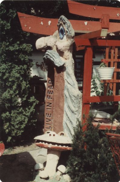 Red painted wooden Japanese torii gate, and a side view of a sculptural arch of a standing old man in Sid Boyum's backyard. The man, with a prodigious nose, big blue eyes, faded ruddy complexion, has a gray toga, and is resting his chin and moustache on an brown painted arch inscribed with the text: "Live In Fear." The "Smiling Mushroom" sculpture is located at the base of the arch. A potted plant is hanging from one of the torii's brackets. 