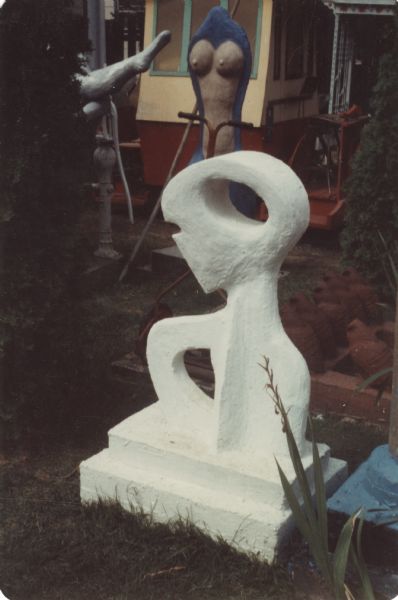 Shaded sculpture, identified as "Creature Abstract," in Sid Boyum's backyard. Painted entirely white, the sculpture uses bold negative spaces with curvilinear shapes to define an abstract human figure: a small head with ocular and a nasal or mouth cutout is sitting on an upper torso, defined by shoulders and open arm or breast. The body stands on a two-tiered base, and in its entirety measures 47" x 31" x 20." In the background are the Toonerville Trolley car, the "Feminine Torso" sculpture and a high-kicking sculptural leg behind a shrub which is near the white hand pump and a push mower. 
