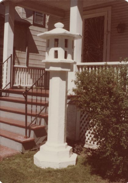 Asian-styled, cast-concrete sculpture identified as "Tall White Lantern" in Sid Boyum's front yard, standing by the steps leading up to the front porch. The lantern has eight sides with sixteen rectangular windows, and is mounted on an octagonal column and topped by an octagonal lid. The entire work measures 87" x 31" x 30."