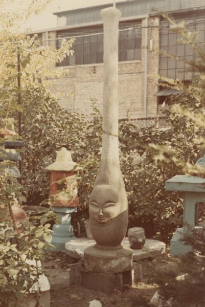 Six-foot high, concrete-cast slender abstract sculpture of an imaginative being with a human face. Jutting out of the head is a narrowing pillar. The eyes are closed with long arched lids, and the nose and mouth are small. It has a round beveled base. It is standing in Sid Boyum's backyard amidst other sculptures along a fence that is adjacent to the Madison-Kipp Corporation.