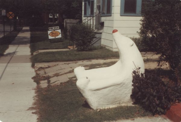 Freshly painted "Polar Bear Chair" sculpture installed in Sid Boyum's front lawn by a driveway and sidewalk and the steps to the front porch of the house. It measures  44" x 40" x 47." On a broad base, its arcing body forms a bucket-like arm chair, with red nose and paws pointing upwards. On the far left is Waubesa Street and a railroad crossing sign.