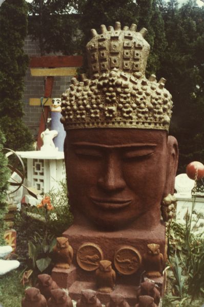 Slightly angled front view of a large, concrete-cast "Monumental Oriental Head" of the Buddha painted red with heavily textured light-colored hair and topknot. Standing 110" x 45" x 78," the sculpture is sitting on a thick base ornamented with Zodiac medallions in the backyard of Sid Boyum's house. The Zodiac symbols in front are of Leo and Scorpio. Sculpted owls are sitting beneath the bust on the base and in the space in front. Part of a white lantern, torii gate, and part of a metal "astrolabe" are pictured to the left rear. The brick building of the Madison-Kipp Corporation is in the far background.