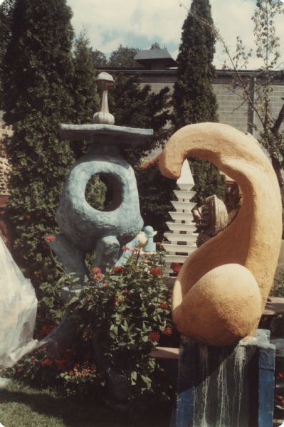 Orange sculpture identified as "Biomorphic Form with Anchor" in Sid Boyum's backyard. It is standing on a blue rectangular base. Also pictured are the "Blue Tripod" and a white pagoda. The brick building of the Madison-Kipp Corporation is in the background. 