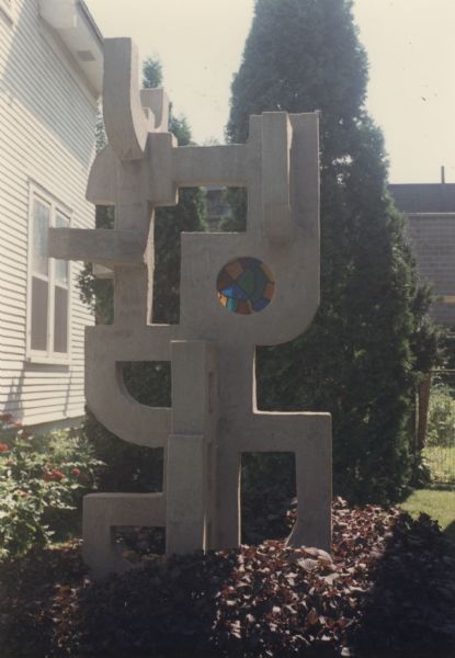 Towering abstract sculpture of cast concrete mortar on a square base, identified as "Geometric with Round Window," near the side of a house. It is composed of intersecting shapes and negative spaces with rounded and squared corners on all four sides, and stands 118" x 62" x 58." The focal point of the sculpture is a round stained-glass window that contains caulk and mortar. The brick building of the Madison-Kipp Corporation is in the background.