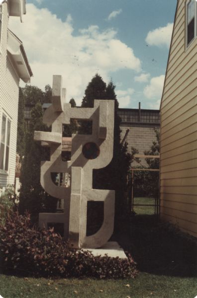 Towering abstract sculpture of cast concrete mortar on a square base, identified as "Geometric with Round Window," near the side of a house. It is composed of intersecting shapes and negative spaces with rounded and squared corners on all four sides, and stands 118" x 62" x 58." The focal point of the sculpture is a round stained-glass window that contains caulk and mortar. The brick building of the Madison-Kipp Corporation is in the background.