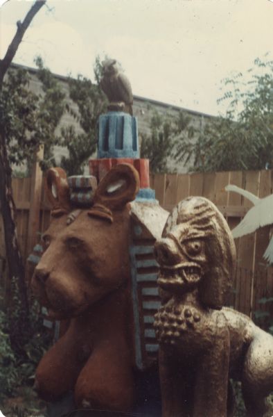 Finished concrete-cast sculpture, "Sphinx," in Sid Boyum's backyard. It's body is brown and it has an Egyptian Pharaoh-styled headdress with blue stripes, and crowned with an owl. The Southeast Asian-styled "Guardian Lion" painted gold is standing next to it in the right foreground. The brick wall of the Madison-Kipp Corporation is in the background beyond the wooden fence. 