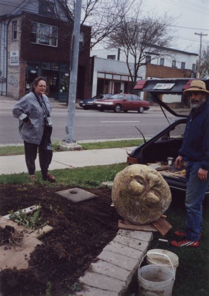 Relocation of Sid Boyum's sculpture, "Smiling Mushroom," to the public green space on the bike path at Atwood Avenue and Dunning Street. Lou Host-Jablonski, AIA architect, is seen at the tail end of a hatchback car in his brimmed hat, and the piece is laying on its side by a newly poured, square concrete foundation for installation. Gretta Wing Miller, producer of several films on the Sid Boyum relocation effort, observes by the sidewalk with her videocamera.   Designated for a "test move" because of its small size, "Smiling Mushroom" was the first piece to be moved from Boyum's backyard for public installation. It was part of a community-based volunteer project to preserve Boyum's larger outdoor work to public parks, landscaped areas and private residences throughout the Atwood-Schenk neighborhood.