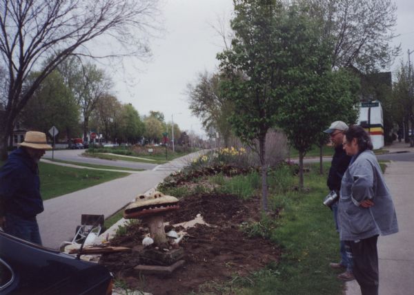 Relocation of Sid Boyum's sculpture, "Smiling Mushroom," to the public green space on the bike path at Atwood Avenue and Dunning Street. Lou Host-Jablonski, AIA architect in the brimmed hat, is seen examining the upright piece standing on a square, concrete foundation for installation. Gretta Wing Miller, producer of several films on the Sid Boyum relocation effort, and Anton ("Tony") Rajer, professional art conservator who specialized in the preservation of folk art, observe by the sidewalk. Spring daffodils are in the background along the bike path.   Designated for a "test move" because of its small size,  "Smiling Mushroom"  was the first piece to be moved from Boyum's backyard for public installation. It was part of a community-based volunteer project to preserve Boyum's larger outdoor work to public parks, landscaped areas and private residences throughout the Atwood-Schenk neighborhood.
