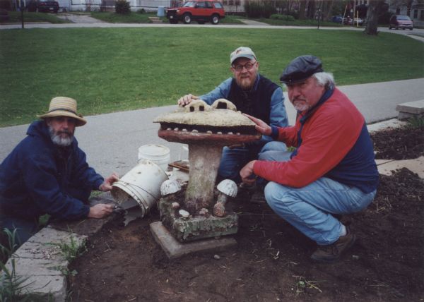 Successful relocation of Sid Boyum's sculpture, "Smiling Mushroom," to the public green space on the bike path at Atwood Avenue and Dunning Street. Lou Host-Jablonski, AIA architect, is seen squatting beside the installed piece, posing with two white plastic buckets and a hand trowel for mixing foundation cement. He is across from Anton ("Tony") Rajer, professional art conservator who specialized in the preservation of folk art, and Steve Boyum (Sid's son and beneficiary), who initiated a preliminary sculptural survey as part of the Save Outdoor Sculpture (SOS) in 1992 with students from the University of Wisconsin-Madison.  Designated for a "test move" because of its small size, "Smiling Mushroom" was the first piece to be moved from Sid Boyum's backyard for public installation. It was part of a community-based volunteer project to preserve Boyum's larger outdoor work to public parks, landscaped areas and private residences throughout the Atwood-Schenk neighborhood.  The "Smiling Mushroom" has a yellow face, red lips, an open mouth with lower teeth, nose and eyes. A few toadstools ring the base of its stem.
