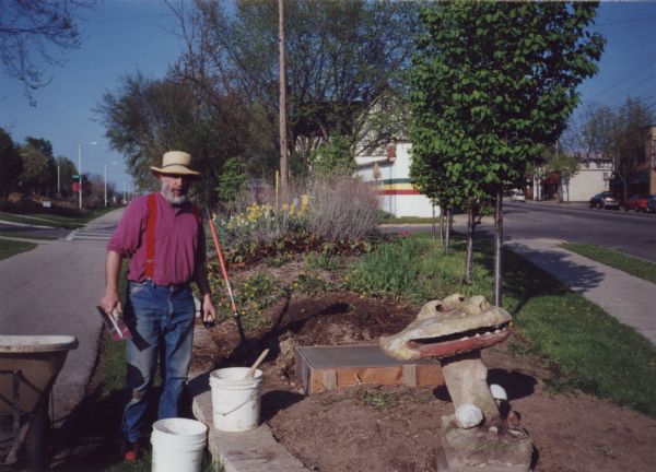 Lou Host-Jablonski, AIA architect, preparing the new concrete foundation for the "Blue Dragon Urn," which was relocated from Sid Boyum's backyard to the public green space on the bike path at Atwood Avenue and Dunning Street. Lou is holding a hand trowel and standing by two white plastic buckets and a shovel stuck in soil. The freshly installed "Smiling Mushroom" sculpture, the first to be moved from Boyum's home, is in front of the new square foundation. Daffodils are in bloom in the background.   The new foundation was part of a major relocation and public installation project that was community-based and volunteer to preserve Boyum's larger outdoor work to public parks, landscaped areas and private residences throughout the Atwood-Schenk neighborhood.
